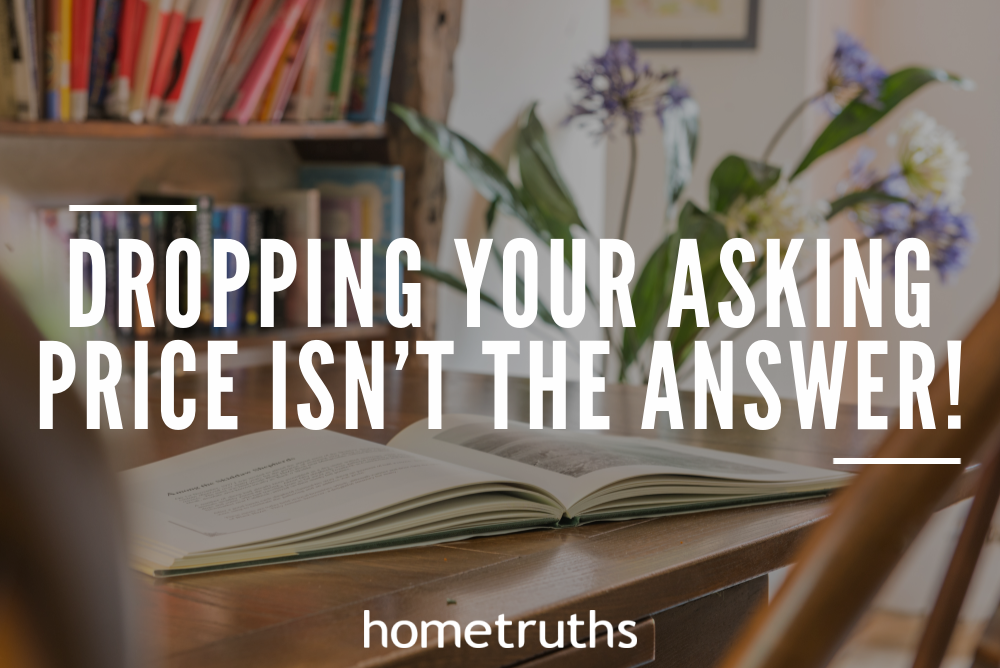 An opened book on top of a wooden table with wooden chairs in front of a bookshelf and a vase with flowers beside it.